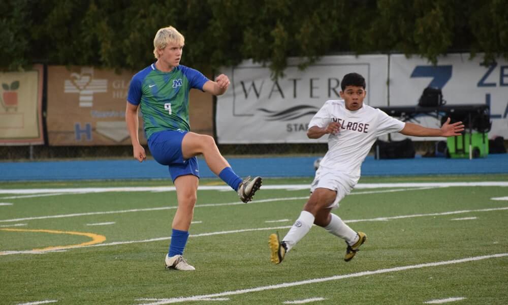 A blond teenager kicking the soccer ball against an opponent